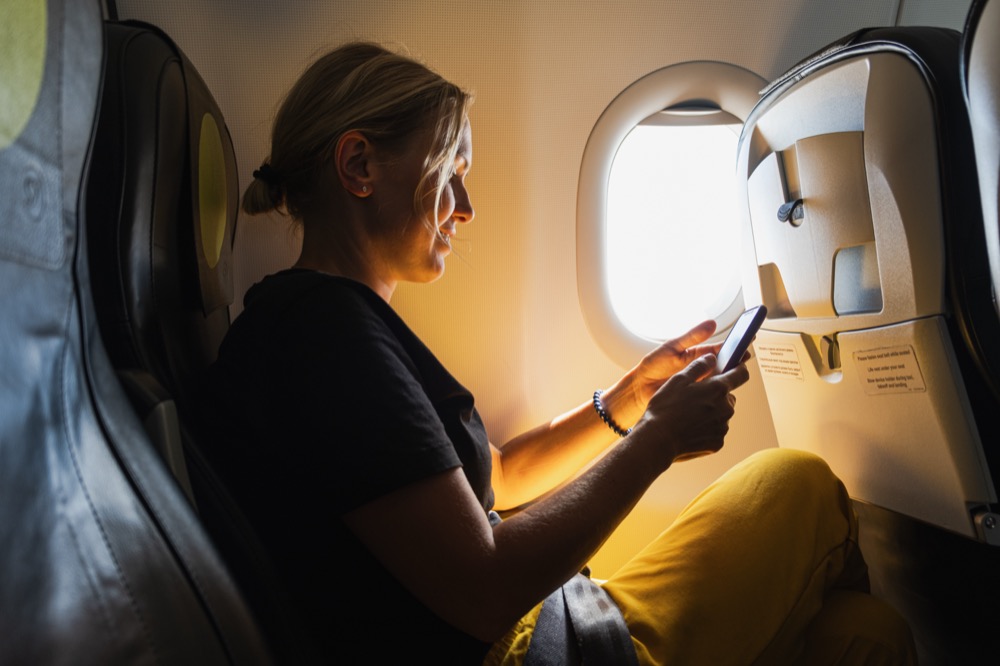 Woman sitting by an airplane window using a smartphone in the sunlight