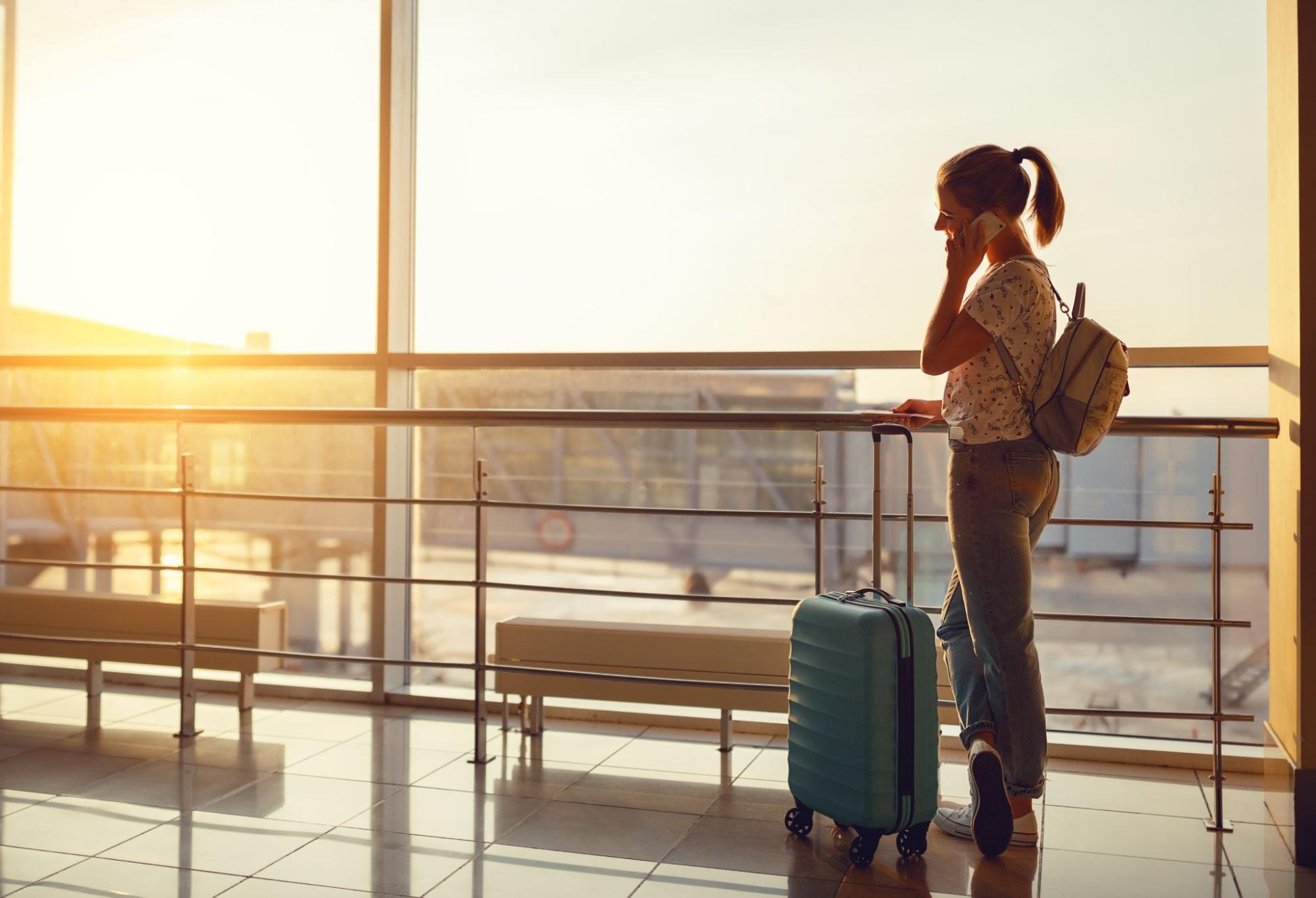 Woman with suitcase and backpack talking on phone at a sunny airport terminal